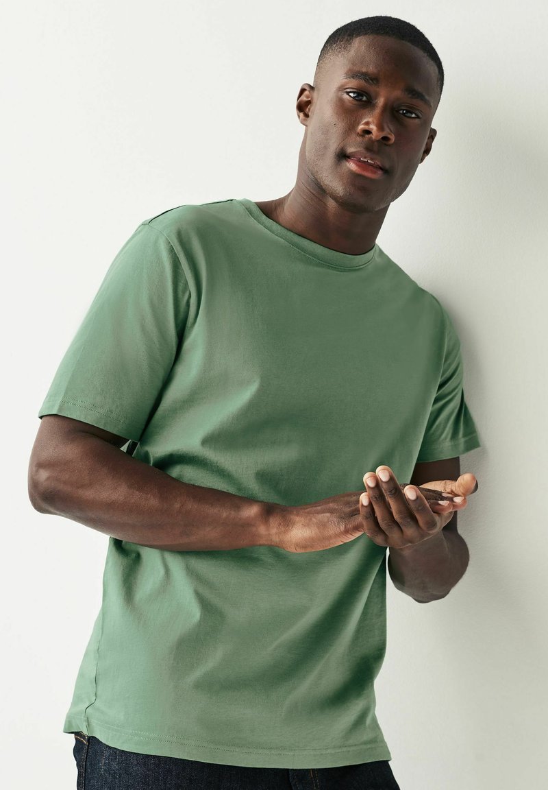 Camiseta de algodón verde con escote redondo y mangas cortas, de textura suave, combinada con jeans oscuros, manos juntas al frente.