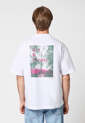 Person mit kurzen braunen Haaren, trägt ein weißes T-Shirt mit grünem und pinkem Grafikdruck mit der Aufschrift "Art & Forest" und blaue Jeans, von hinten gesehen.