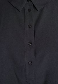 Crna polo majica s klasičnom ovratnikom, s plaketom koja ima pet crnih dugmadi i glatkom teksturom. Jednostavan i čist dizajn.