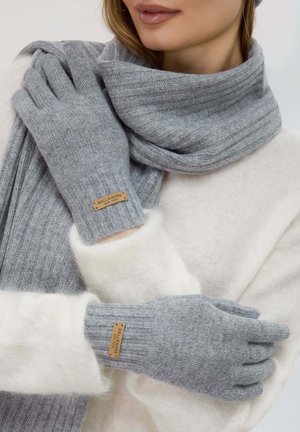 Guanti grigi lavorati a maglia con texture a coste e etichetta in pelle; abbinati a una sciarpa grigia coordinata. Lo sfondo presenta un morbido maglione bianco.