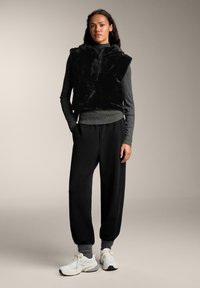 Vesta neagră, asemănătoare cu blana, cu glugă, peste un pulover gri cu guler înalt. Pantaloni negri, tapered, cu manșete elastice. Adidași albi cu accente gri.