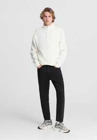 Pullover bianco con colletto alto e zip, abbinato a pantaloni neri affusolati e sneaker grigie, in piedi contro uno sfondo chiaro.