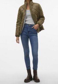 Chaqueta acolchada verde oliva con cuello elevado, combinada con jeans azul ajustados y botines marrones, que exhibe una silueta moderna y elegante.
