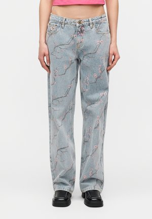 Άτομο που φοράει τζιν παντελόνι ανοιχτού μπλε με ροζ λουλουδάτα σχέδια σε κλαδιά, μαύρα παπούτσια και ένα γκριμμένο ροζ τοπ, σε απλό φόντο.