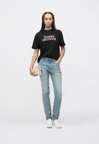 Μαύρο κοντομάνικο T-shirt με το λογότυπο "TOMMY HILFIGER", ανοιχτό μπλε τζιν παντελόνι στενής εφαρμογής, λευκά αθλητικά παπούτσια και μπεζ τσάντα.