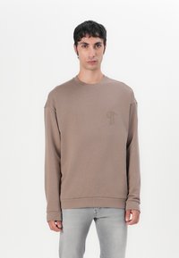 Sudadera marrón de tejido suave, con un cuello redondo y mangas largas. Incluye un sutil logo en relieve en el lado izquierdo del pecho.