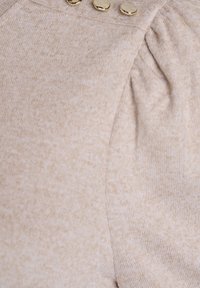 Béžová pletená látka s jemnou texturou a zlatými knoflíky v horní části. Design zahrnuje nařasené detaily poblíž ramenní oblasti.