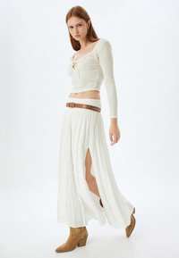 Witte, lange gelaagde rok met een hoge split tot boven de dij, gecombineerd met een fitted, gestructureerd wit top met lange mouwen en een strikdetail. Bruine riem.