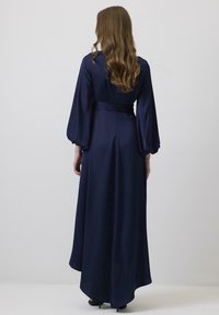 Rochie lungă din satin de culoare albastru închis, cu mâneci ample și un talie legată. Textură netedă, cu tiv până la podea și o trenă subtilă.