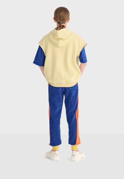 Personne aux cheveux attachés, vue de dos, portant un gilet jaune sans manches à capuche, une chemise bleue, un pantalon bleu avec des rayures orange et des chaussures blanches.