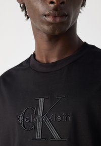 Krupan plan osobe koja nosi crnu majicu s reljefnim Calvins Klein logotipom centriranim na prsima, na jednobojnoj pozadini.