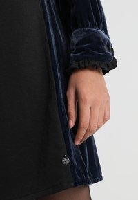 Hand in natürlichem Hautton, die neben einem langen dunkelblauen Samtkleid mit schwarzen Stoff- und Rüschendetails am Ärmel ruht.