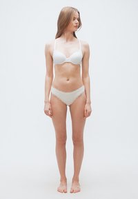Νεαρή γυναίκα όρθια, ξυπόλυτη, φορώντας λευκό σουτιέν και ταιριαστά εσώρουχα με δαντέλα, μπροστά σε απλό λευκό φόντο.