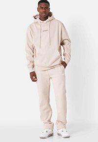 Felpa cappuccio beige e pantaloni felpati realizzati in tessuto morbido, con tasca a canguro, stringhe e sottili dettagli con logo, abbinati a sneakers bianche.