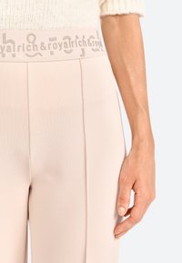 Pantaloni rosa chiaro con tessuto texture e una cintura ampia con dettaglio logo embossato, mostrati con una mano sulla tasca laterale.