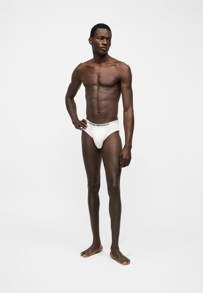 Λεπτός, σκουρόχρωμος άνδρας στεκόμενος ξυπόλυτος, φοράει λευκά εσώρουχα Emporio Armani, με το χέρι στη μέση, κοιτάζοντας αριστερά σε απλό λευκό φόντο.