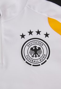 Λευκό αθλητικό �μπουφάν με το λογότυπο του Γερμανικού Συνδέσμου Ποδοσφαίρου, μαύρο έμβλημα αετού και τέσσερις αστέρες από πάνω. Συμπεριλαμβάνει μαύρες και κίτρινες λεπτομέρειες.