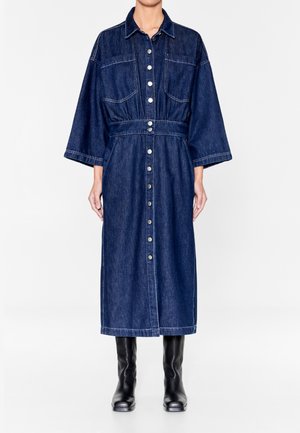Robe longue en denim foncé boutonnée, manches larges et taille ceinturée, portée avec des bottes noires montantes jusqu'aux genoux.