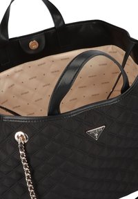 Czarna pikowana torba typu tote z czarnymi skórzanymi uchwytami, wnętrze z wzorzystą beżową podszewką oraz srebrne logo GUESS. Torbę zdobi detal z łańcuszka.