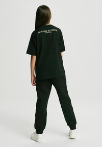 Czarny, oversize t-shirt z białym napisem "DIVERSE EXTREME" na plecach, zestawiony z pasującymi czarnymi spodniami i białymi trampkami. Gładka tekstura materiału.