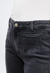 Κοντινή λήψη μαύρου τζιν με πίσω τσέπη και ορειχάλκινο κουμπί, φορεμένο με λευκό ριμπ τοπ.