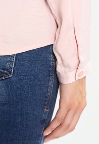 Ljust rosa långärmad blus med rund krage och knäppning vid ärmsluten, i kombination med mörkblå jeans i denim. Slät tygstruktur.