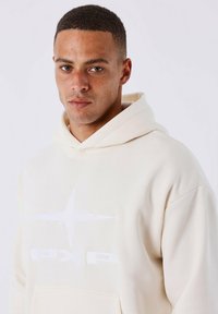 Licht beige hoodie van zachte stof, met een voorzak en een wit logo met een sterdesign. Aangehechte capuchon en een relaxte pasvorm.