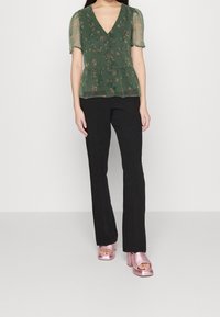 Blouse fleurie verte avec manches en voile, col en V et design peplum, associée à un pantalon noir évasé et des chaussures à plateforme roses brillantes.