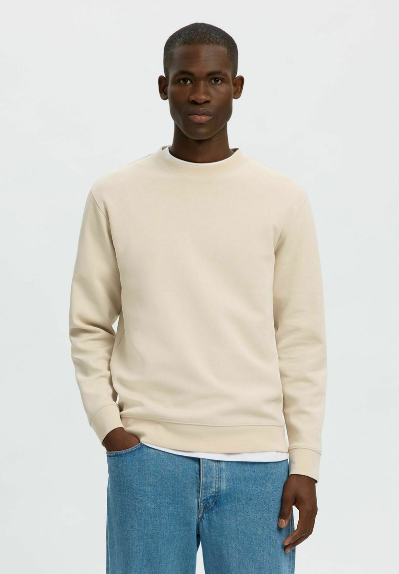Jeune homme à la peau foncée portant un sweat-shirt beige et un jean bleu, debout avec une main dans la poche devant un fond uni.