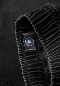 Juodos medžiagos detalė su surinkta dalimi, kurioje pritvirtintas mažas skaidrus mygtukas prie pereinamo ribbingo dirželio. Tekstūra lygi su subtiliais siūlais.