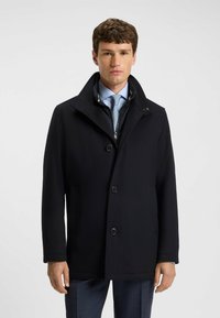 Mann in einem dunkelblauen Mantel über einem blauen Hemd, mit einer hellblauen Krawatte und dunkel gemusterten Hosen, der vor einem einfarbigen hellen Hintergrund steht.