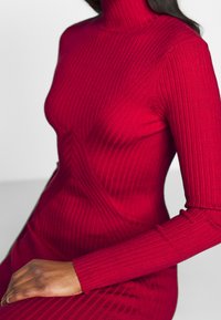 Red ribbed turtleneck dress with long sleeves, featuring a fitted silhouette and diagonal pattern detailing. Soft, textured fabric.