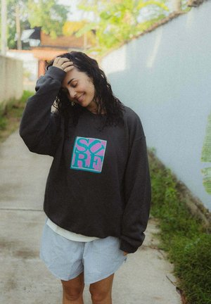 Czarna, oversize'owa bluza z kolorowym napisem "SURF", zestawiona z jasnoniebieskimi, pasiastymi szortami. Ubranie noszone na zewnątrz.