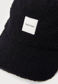 Fekete, textúrált polársapka lapos szélekkel és fehér feliratozott címkével, amelyen a "Calvin Klein" felirat szerepel. Puha, meleg anyag és lekerekített forma.