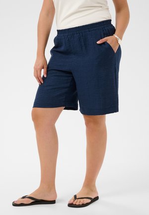Person trägt lockere marineblaue Shorts, weißes Top, schwarze Flip-Flops und ein silbernes Armband, steht mit einer Hand in der Hosentasche.