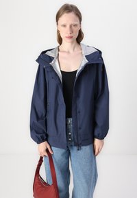 Giacca leggera blu navy con cappuccio, polsini elasticizzati e fodera interna argentata. Abbinata a jeans a vita alta e una borsa rossa.