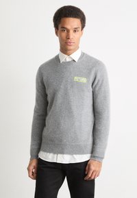 Pilkas vilnos megztinis su gumuotomis rankovėmis ir apačia. Ant krūtinės išsiskiria žalias "FUTURE" logotipas. Dėvimas ant baltos marškinių su apykakle.