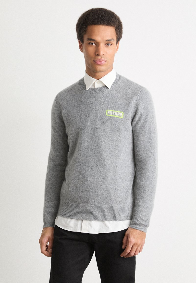 Pilkas vilnos megztinis su gumuotomis rankovėmis ir apačia. Ant krūtinės išsiskiria žalias "FUTURE" logotipas. Dėvimas ant baltos marškinių su apykakle.