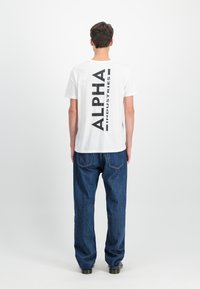 Balts kokvilnas T-krekls ar lielu melnu "ALPHA INDUSTRIES" tekstu, kas vertikāli izvietots aizmugurē, kombinēts ar zilu džinsu biksēm.