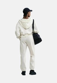 Cremfarbener Kapuzenpullover und passende tapered Sweatpants, kombiniert mit einer schwarzen Tasche und Sneakers, von hinten vor einem schlichten Hintergrund betrachtet.