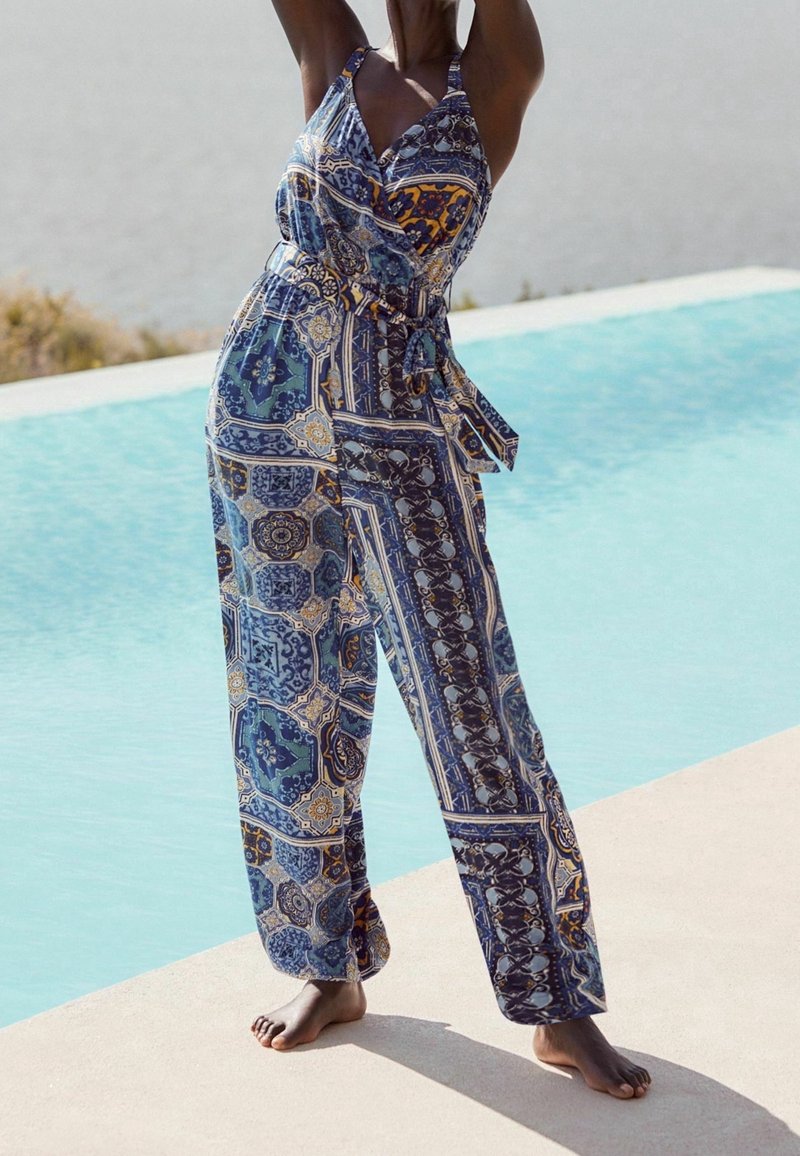 Femme portant une combinaison sans manches à motifs, debout pieds nus près d'une piscine, les bras levés au-dessus de sa tête.