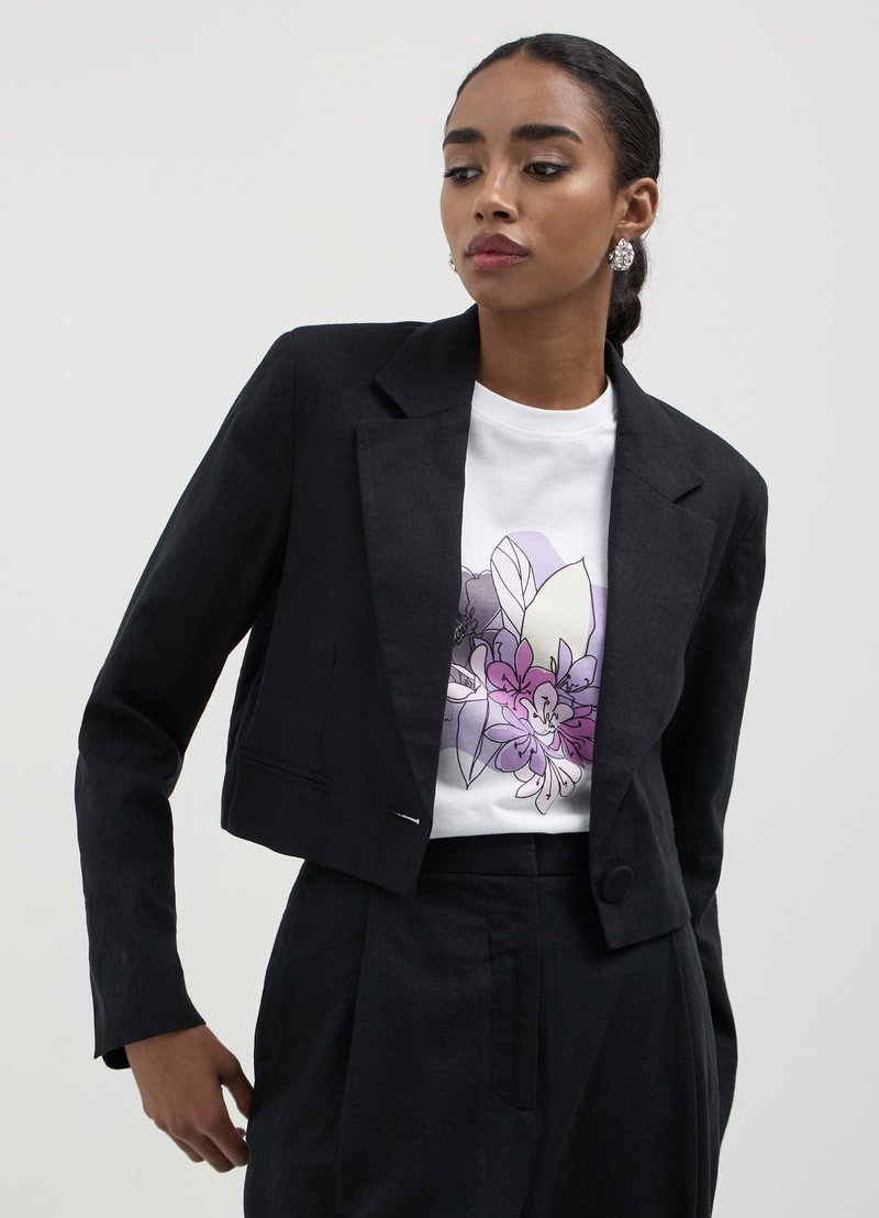 Blazer noir court avec col cranté et fermeture à bouton, associé à un t-shirt blanc avec un design graphique floral coloré.