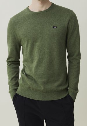 Homme portant un pull ras du cou vert olive avec un petit logo rectangulaire noir et blanc sur la poitrine, associé à un pantalon noir.