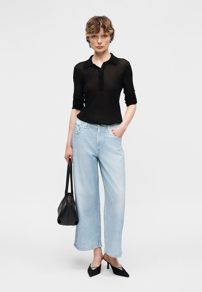 Γυναίκα που φοράει μαύρο πόλο μπλούζα με μακριά μανίκια, ανοιχτό μπλε παντελόνι φαρδιάς γραμμής, μαύρες γόβες και κρατάει μια μαύρη τσάντα χειρός, στέκεται μπροστά σε ένα απλό φόντο.