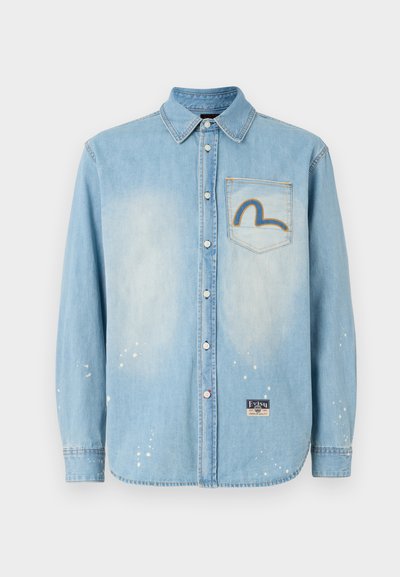 Camicia di jeans azzurro chiaro con maniche lunghe, chiusura a bottoni, dettagli strappati e un design arcobaleno ricamato sul taschino sinistro.