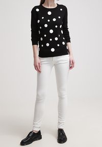 Czarny sweter w białe grochy, z długimi rękawami i okrągłym dekoltem. Noszony z białymi dopasowanymi jeansami i czarnymi sznurowanymi butami.