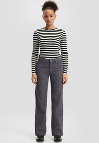 Woman with curly hair wearing a black and white striped long-sleeve top, gray corduroy pants, and black shoes, standing against a plain background.
