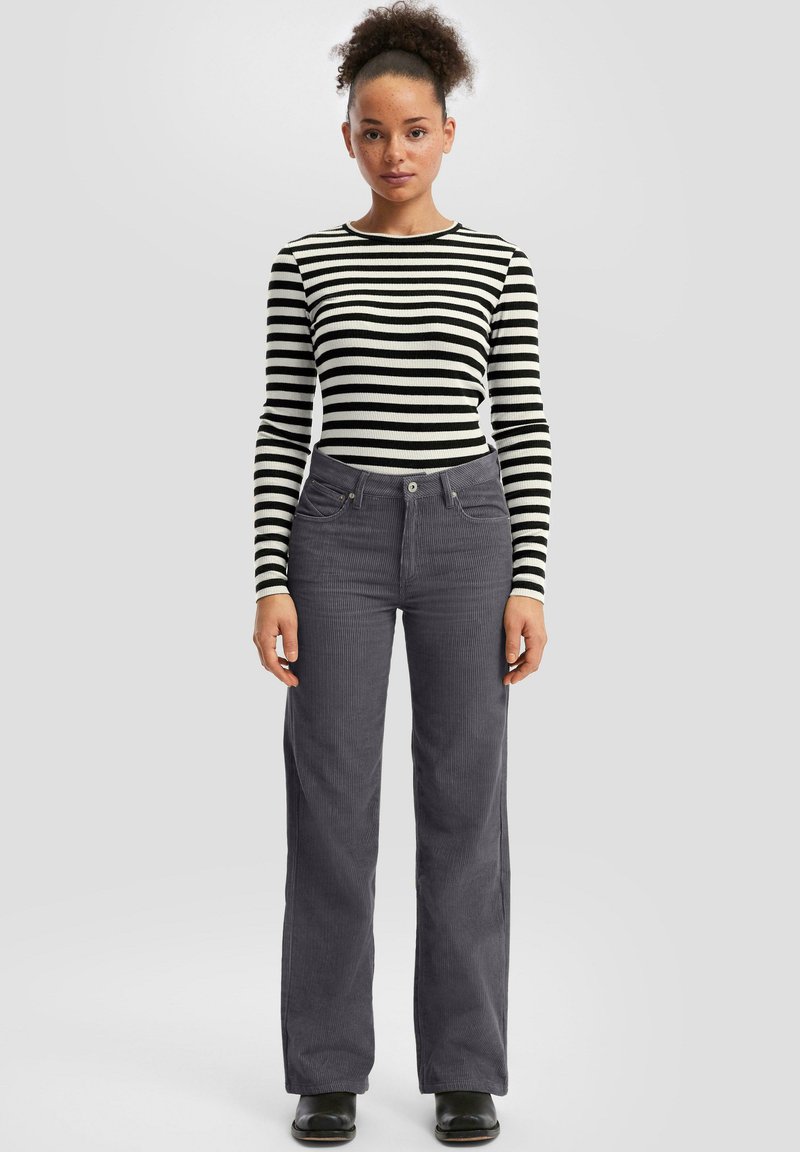 Woman with curly hair wearing a black and white striped long-sleeve top, gray corduroy pants, and black shoes, standing against a plain background.