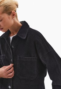 Czarna kurtka z korduroy z kołnierzem, dużą przednią kieszenią i zapięciem na guziki, z pionowymi żebrami i luźnym krojem.