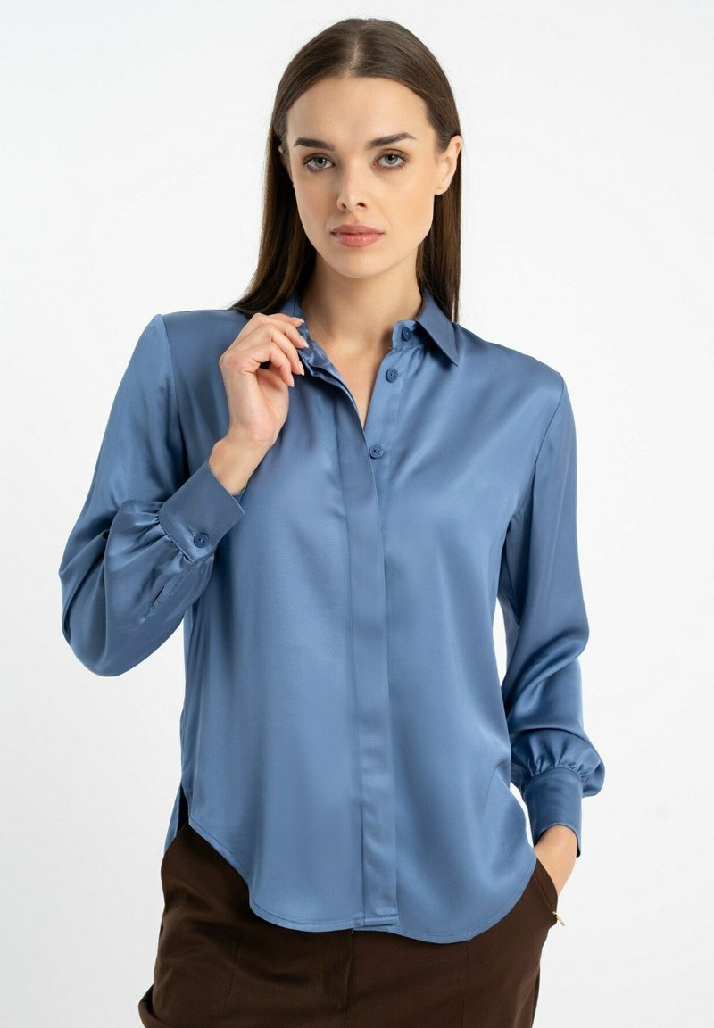 Saratynowa niebieska koszula z długim rękawem, z kołnierzykiem zapinanym na guziki, marszczonymi mankietami i zaokrąglonym dołem, w połączeniu z ciemnobrązowymi spodniami.